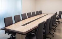 A modern boardroom before a meeting: a long boat-shaped table of pale figured maple with a dark border, every chair tucked in along both sides, a single glass of water at the head, and one chair standing just past the end of the table, turned back toward it
