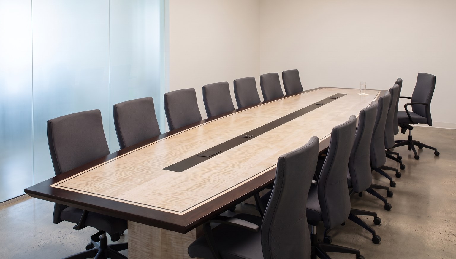 A modern boardroom before a meeting: a long boat-shaped table of pale figured maple with a dark border, every chair tucked in along both sides, a single glass of water at the head, and one chair standing just past the end of the table, turned back toward it