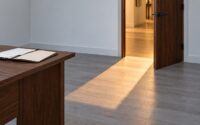 An unbroken band of warm corridor light falling through an open office doorway onto a cool grey wood floor, with the corner of a walnut desk and an open folder at the left of the frame and no one in the doorway