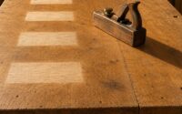 An old oak workbench in warm raking light, marked by a row of pale sun-bleached rectangles where objects once sat, with a single worn wooden hand plane remaining at the right and casting the only shadow in the frame